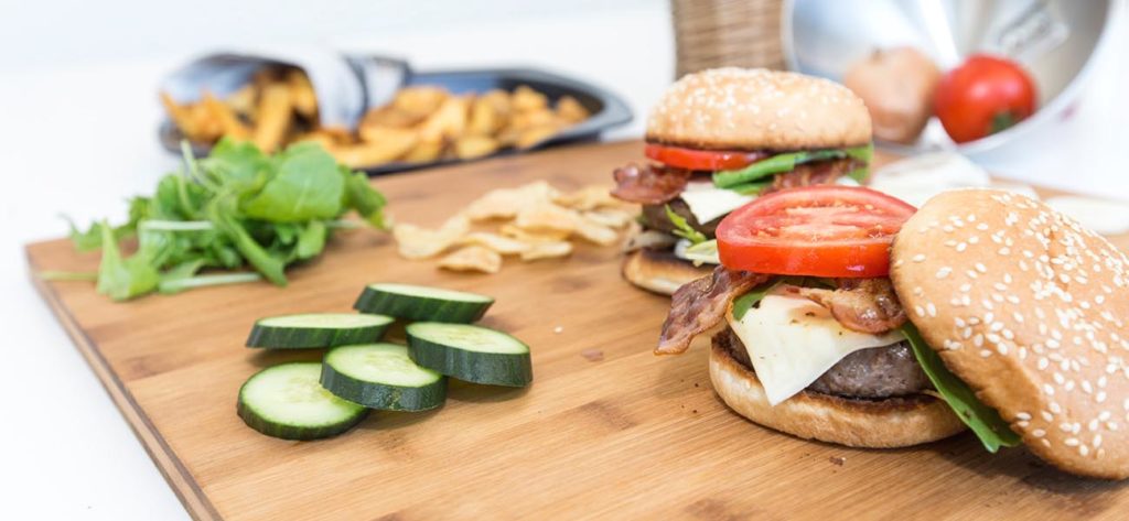 Schneidebrett mit Gurkenscheiben, Rucola-Salat und zwei angerichteten Burgern, in Hintergrund liegen Pommes frites.