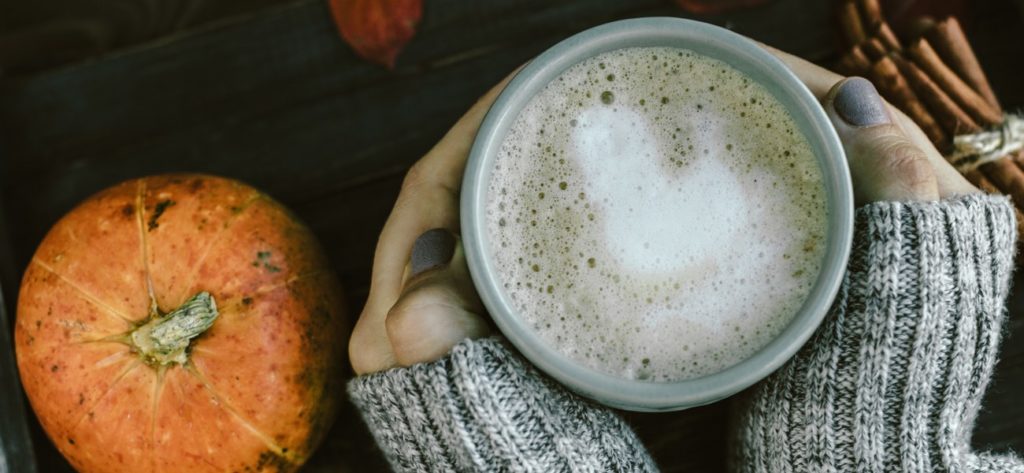 Frauenhände halten einen herbstlichen Kaffee mit Milch neben Kürbis und Zimt.