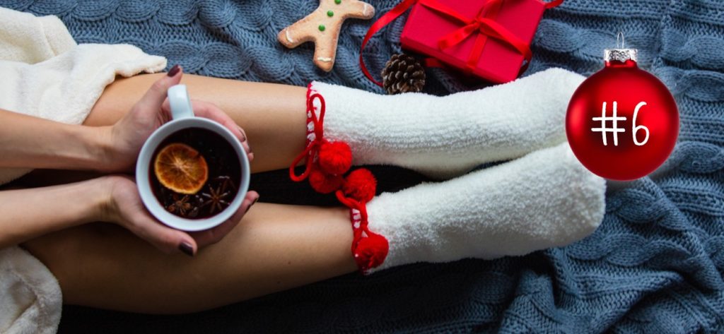 Beine auf einer Wolldecke von oben, Füsse in Wollsocken. Festliche Dekoration auf der Wolldecke, Hände halten eine Tasse mit Weihnachtsgetränk.