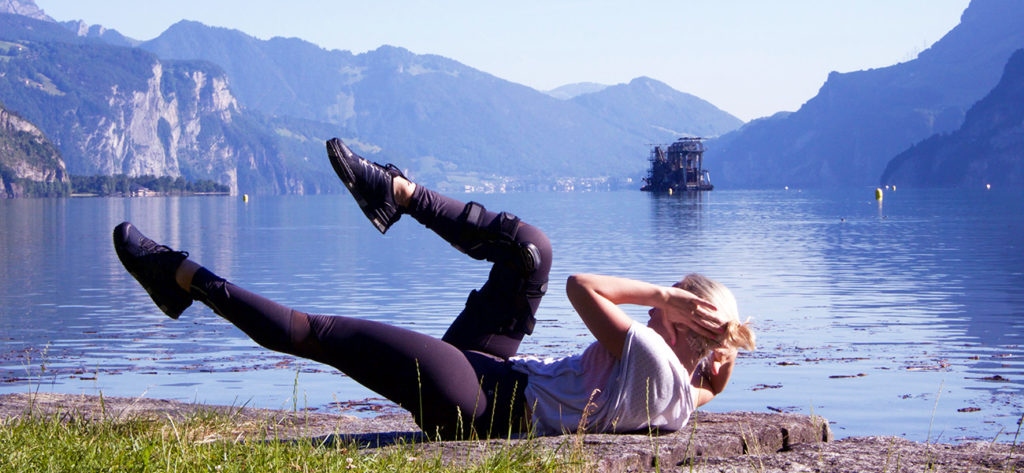 blonde Frau liegt auf grünem Rasen und macht Bauchmuskel Übungen, im Hintergrund hat es einen See und Berge