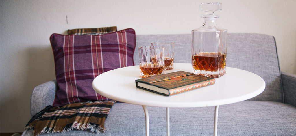 weisses rundes Tischlein mit einem Whisky-Krug und zwei Gläsern drauf, zudem liegt ein blumengemusterter Block auf dem Tisch. Im Hintergrund hat es ein hellgraues Sofa mit einem rot-kariertem Kissen und einem braun-kariertem Schal.